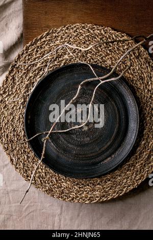 Empty black ceramic plate with dry branch on straw napkin and wooden background. Flat lay, space. Stock Photo