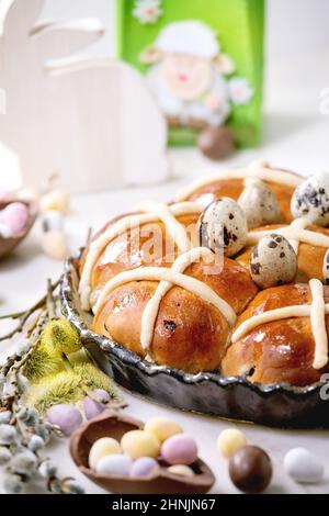 Homemade Easter traditional hot cross buns in ceramic dish with blossom flowers, chocolate candy and quail eggs on white marble table. Easter holiday Stock Photo