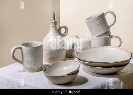 Set of empty craft ceramic tableware, white speckled bowls, plates, mugs and oil bottle standing on white table cloth. Stock Photo