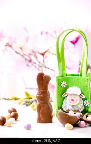 Easter greeting card with chocolate sweets rabbit, green bag with lamb, candies and eggs, pink spring flowers on white background. Easter treats Stock Photo