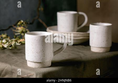 Set of white speckled empty craft ceramic coffee tea mugs standing on dark green linen tablecloth with stack of plates. Stock Photo