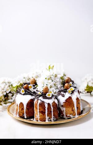 Homemade traditionla Easter kulich cake with chocolate nests and eggs on ceramic plate and blossoming cherry tree over white texture background. Tradi Stock Photo