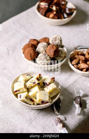 Collection of homemade toffee chocolate almond nuts candy, dark chocolate truffle, white chocolate candy bar in bowls on white linen table cloth. Hand Stock Photo