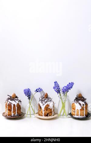 Homemade traditionla small Easter kulich cakes with chocolate nests and eggs on plates in row decorated with muscari flowers over white marble table. Stock Photo