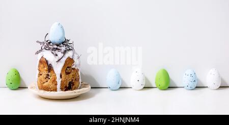 Homemade traditionla Easter kulich cake with chocolate nest, eggs on plate standing on white table with colored eggs in row. Traditional ortodox easte Stock Photo