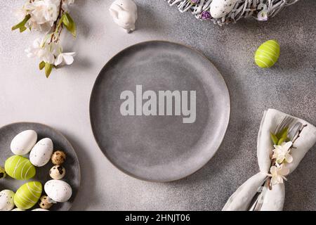 Easter table setting with gray plate, spring blossom flowers and white green colorful eggs on gray background. Top view. Copy space. Stock Photo