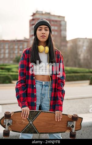 Beautiful young hipster woman posing with skateboard. Hispanic Latin woman wearing casual clothes. Stock Photo