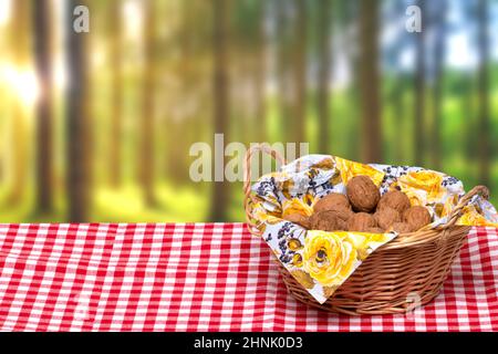 A wicker basket with a napkin full of walnuts on a table with a red checkered tablecloth in front of an abstract blurred natural green forest backgrou Stock Photo