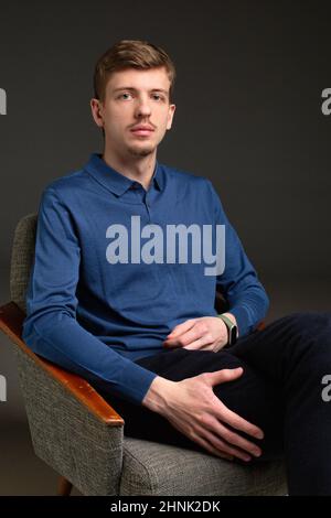 Young blond european man with short beard and mustache isolated on black sitting in armchair Stock Photo