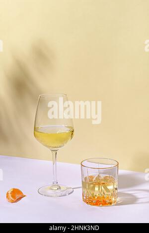 Artistic still life with two different glasses of white wine next to a slice of orange outdoors on a white table against yellow wall for copy space Stock Photo