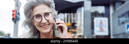 Happy grey haired senior woman with glasses talks on mobile phone on city street Stock Photo
