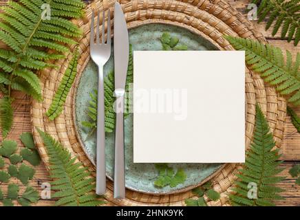 Square blank paper card on table setting decorated with fern leaves on wooden table top view. Tropical mock-up scene with invitation card flat lay Stock Photo