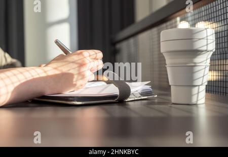 Female hands closeup taking notes in notebook. Woman sitting at table with mug in cafe and planning day or writing thoughts, information in planner. High quality photo Stock Photo
