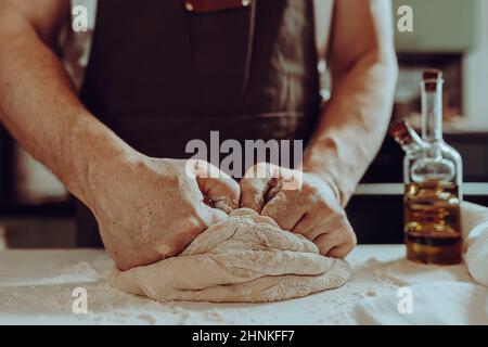 male baker, in brown apron, kneads dough with strong muscular arms, standing in home kitchen. Cooking homemade dough, bread, homemade noodles Stock Photo