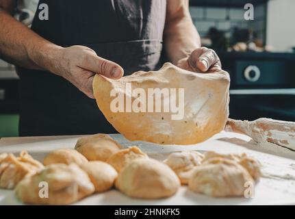 muscular hands of male baker hold thinly rolled dough. Cooking in home kitchen dough for pizza or pita bread. man prepares homemade cakes Stock Photo