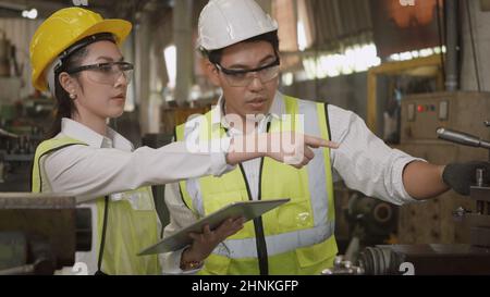 mechanical engineer woman and operation man wearing uniform hardhat and goggles safety working on workshop metal lathe Stock Photo