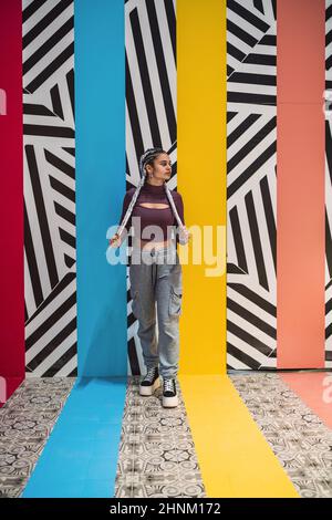 Young woman standing clutching her braids while looking away against wall with colors and striped designs Stock Photo