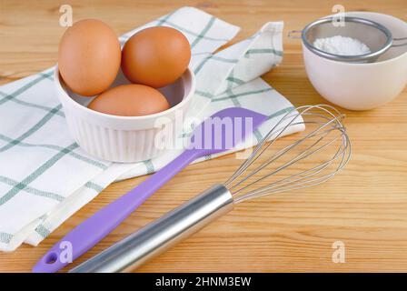 A whisk and spatula with eggs and kitchen utensils on wooden table Stock Photo