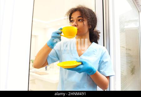 Young african american female doctor in uniform and gloves drinking coffee or tea while taking break between patient consultations in clinic, medical Stock Photo