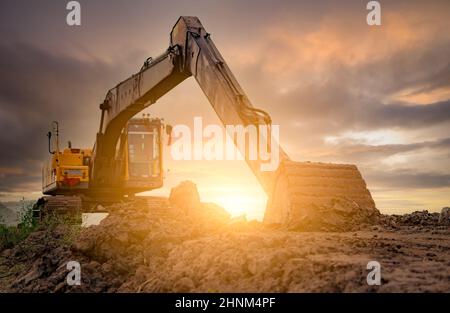 Backhoe parked at construction site after digging soil. Bulldozer on sunset sky and clouds background. Digger after work. Earth moving machine at construction site at dusk. Digger with dirt bucket. Stock Photo