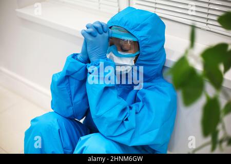 Tired female doctor in personal protective equipment PPE feeling depressed after difficult stressful shift during Covid-19 pandemic, sitting on floor Stock Photo