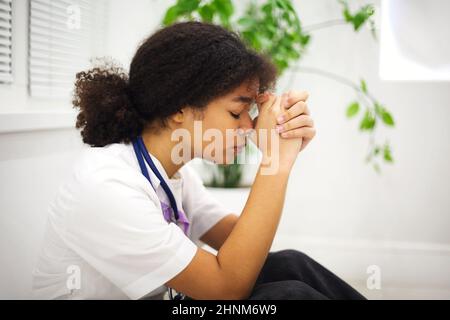 Sad depressed young african american nurse sitting on floor with frustrated face expression, feeling overwhelmed by workload in clinic, ethnic female Stock Photo