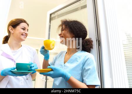 Two happy multi-ethnic female medical workers colleagues drinking coffee or tea and talking while standing near window in modern clinic or hospital, t Stock Photo