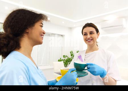Two happy multi-ethnic female medical workers colleagues drinking coffee or tea and talking while standing near window in modern clinic or hospital, t Stock Photo