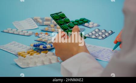Doctor hand writing a prescription holding a pill blister. Pile of pills in blister packs on blue background. Stock Photo