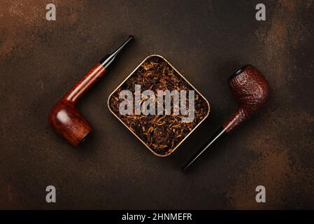 Close up tin full of ready rubbed long coarse cut tobacco blend and two handmade briar smoking pipes over grunge brown background, elevated top view, Stock Photo