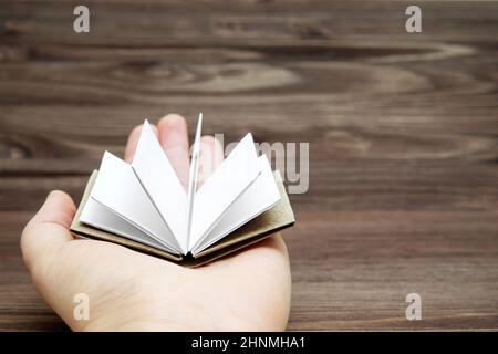 Accordion folded open miniature book with blank pages on the palm of the female hand against a wooden table background. Education and reading concept. Stock Photo