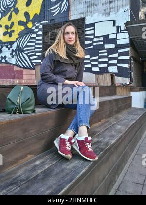 A young woman in a coat, jeans and sneakers sits on the wooden steps of a painted wall Stock Photo