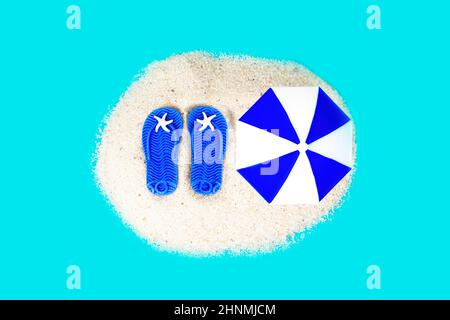 Miniature lip flops and sun umbrella on a pile of sand isolated on a blue background. Creative sandy beach vacations concept. Stock Photo