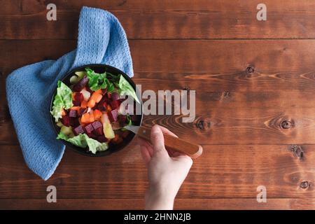 Woman hand eating salad of beet, carrot and lettuce in bowl on wooden background. Copy space. Top view. Stock Photo