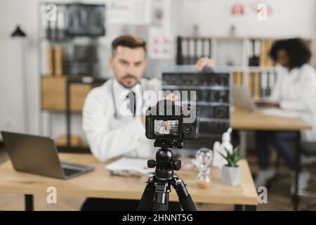 Competent male physician in uniform recording video blog while examining x ray scan at hospital room. Focus on digital camera fixed on tripod. Stock Photo