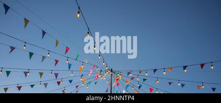 Garland with colorful pennants and light bulbs on pole over blue sky. Long format Stock Photo