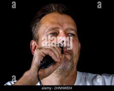A middle-aged man cuts his gray beard with a shaving trimmer. Stock Photo