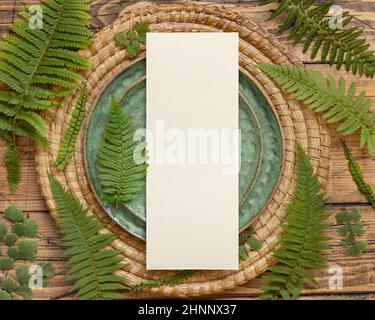 Blank paper card laying on green plate on brown wooden table with fern leaves top view. Tropical mock-up scene with invitation card flat lay Stock Photo
