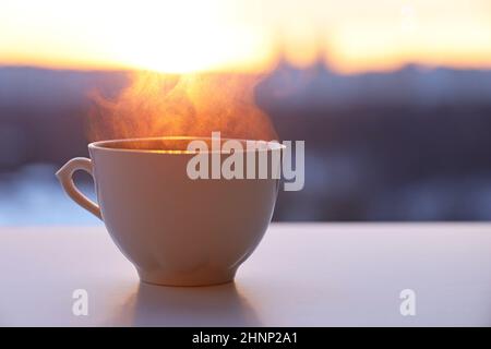 Hot coffee or tea cup with smoke on sunshine background. View from the window to silhouettes of city buildings Stock Photo