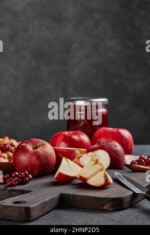 Still life with fresh lingonberries, red appples with knife on black cutting board and jar of homemade jam on dark background with copy space. Autumn Stock Photo