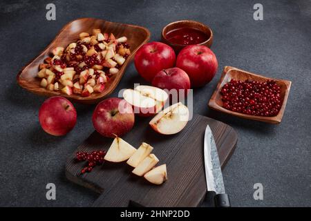 Still life with fresh lingonberries, red appples with knife on black cutting board and  homemade jam on wooden bowl on dark background. Top view. Autu Stock Photo
