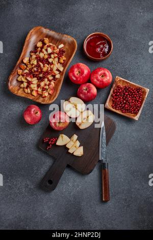 Still life with fresh lingonberries, red appples with knife on black cutting board and  homemade jam on wooden bowl on dark background. Top view. Autu Stock Photo