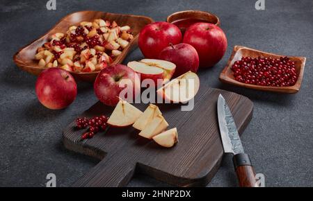 Still life with fresh lingonberries, red appples with knife on black cutting board and  homemade jam on wooden bowl on dark background. Top view. Autu Stock Photo