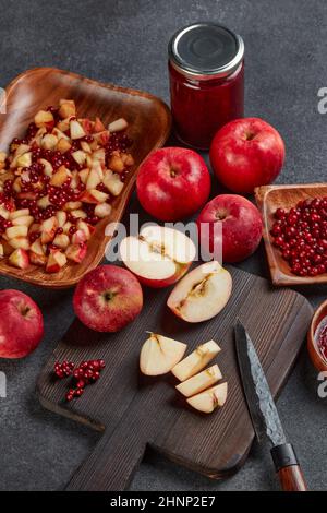 Still life with fresh lingonberries, red appples with knife on black cutting board and  homemade jam on wooden bowl on dark background. Top view. Autu Stock Photo
