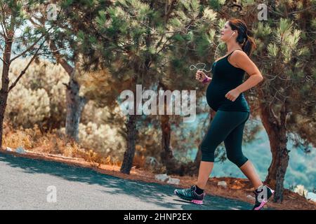 Happy Pregnant Woman on Workout in the Park on Sunny Day. Enjoying Sportive Walk Among Beautiful Fresh Trees. Active Healthy Pregnancy. Stock Photo