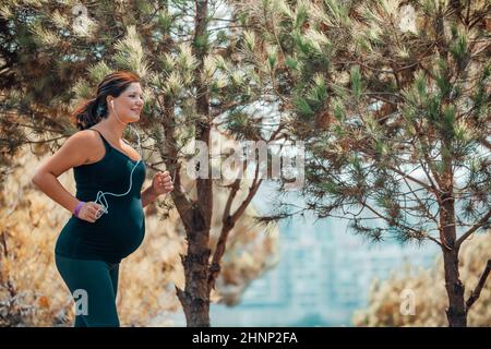 Sportive Pregnant Woman Jogging in the Mountainous Forest on Sunny Day. Enjoying the Walk Among Fresh Pine Trees. Happy Healthy Pregnancy. Stock Photo