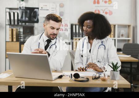 Caucasian male doctor and african american female nurse sitting together at desk, talking and using modern laptop. Medical workers in white lab coats having discussion at hospital room. Stock Photo