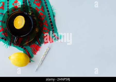 A cup of tea with lemon stands on a dark saucer, which stands on a knitted green-red scarf, next to it lies a thermometer and a lemon on a white backg Stock Photo