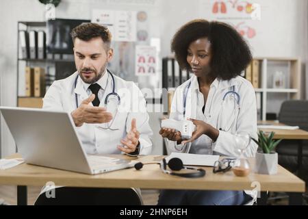 Multiracial male and female doctors using modern laptop for online seminar. Two medical workers sitting at desk and talking about usage of contactless infrared thermometer. Stock Photo