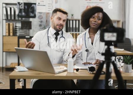 View through video camera on multiracial medical workers sitting at desk in face masks and lab coats. Two competent doctors recording vlog at modern hospital. Stock Photo